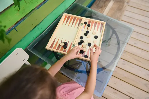 stock image A child engaging in a game of backgammon at a glass table on a vibrant, green and wooden deck, showcasing leisure and strategic thinking.