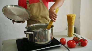 Kırmızı tişörtlü ve önlüklü biri fırında İtalyan makarnası hazırlıyor, tezgahta da taze domates ve spagetti var..