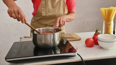 Taze domatesler, spagetti ve bir kase ile çevrili bir tencerede domates sosu karıştıran biri. Ev yapımı İtalyan yemekleri ve taze malzemeler kavramı.