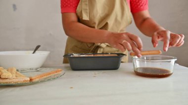 Bir kadının tiramisu yaptığı yakın plan bir fotoğraf, bayan parmak bisküvilerini kahveye bandırma, ve onları tavaya dizme, ev mutfağında klasik bir İtalyan tatlısı hazırlama,.
