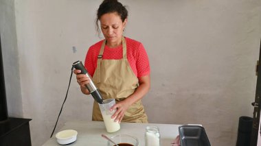 Modern bir mutfakta malzemeleri karıştırmak için el blenderi kullanan bir kadın. İtalyan yemeklerini odaklanma ve hassasiyetle hazırlıyorum..