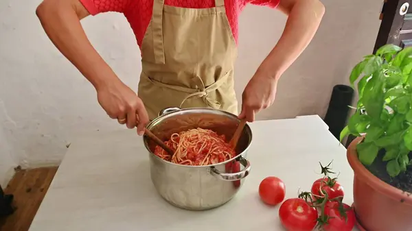 stock image A person in an apron cooking Italian pasta with tomato sauce in a cozy kitchen, featuring fresh tomatoes and a basil plant on the counter.