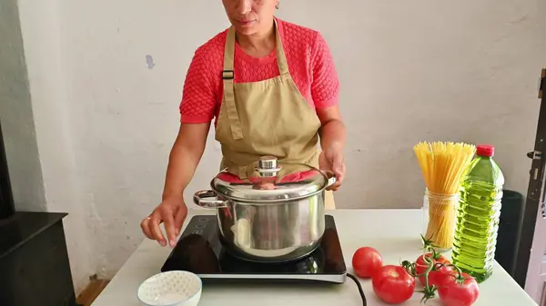 stock image A woman cooking Italian food in her kitchen, preparing a meal with fresh tomatoes, spaghetti, and olive oil. She uses an electric stove and has a pot on the cooktop.