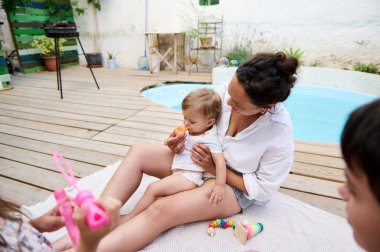 Bir anne bebeğiyle havuz kenarında oturur ve güneşli bir günde çocuklarla köpüklerin tadını çıkarır. Aile eğlencesi ve dışarıda rahatlama..