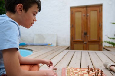 Genç bir çocuk havuzun yanındaki ahşap bir güvertede oturuyor, satranç oynamaya odaklanmış. Parlak ve çekici bir konsantrasyon anı.