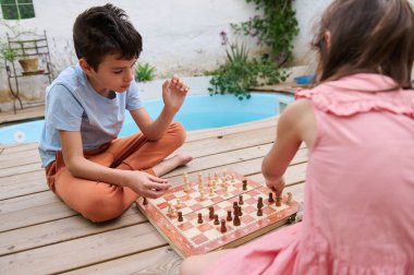 İki çocuk havuzun yanındaki tahta güvertede satranç oynuyorlar. Sahne, entellektüel gelişimi ve açık hava aktivitelerini teşvik eden neşeli ve dingin bir sahne..