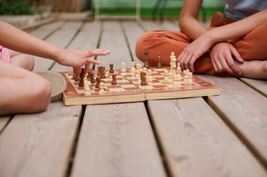 Ahşap bir güvertede satranç oynayan çocukların yakın çekimi stratejik düşünme, arkadaşlık ve açık hava eğlencesini vurguluyor. Hareket eden eller çarpışma ve konsantrasyonu gösterir.