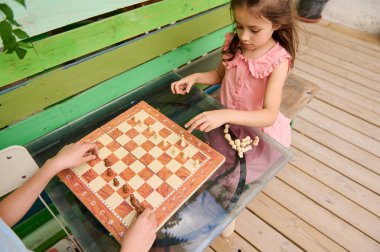 Dışarıda tahta bir masada satranç oynayan iki çocuk. Açık hava ortamında strateji, konsantrasyon ve eğlence vurgulanıyor.