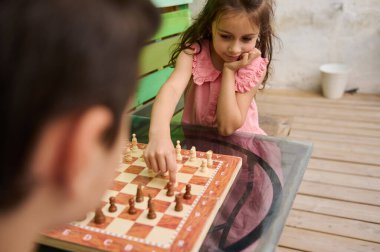 Pembe elbiseli genç bir kız ahşap bir masada arkadaşlarıyla oyun oynarken satranç tahtasına bir parça koyar..