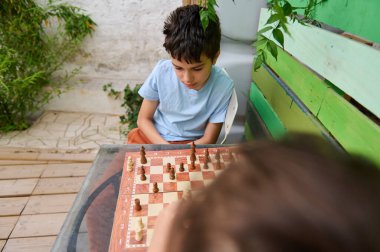 Genç bir çocuk, bir arkadaşıyla açık hava bahçesinde satranç oynamaya yoğun bir şekilde odaklanıyor. Yeşillik ve canlı ahşap bir duvar içeriyor..