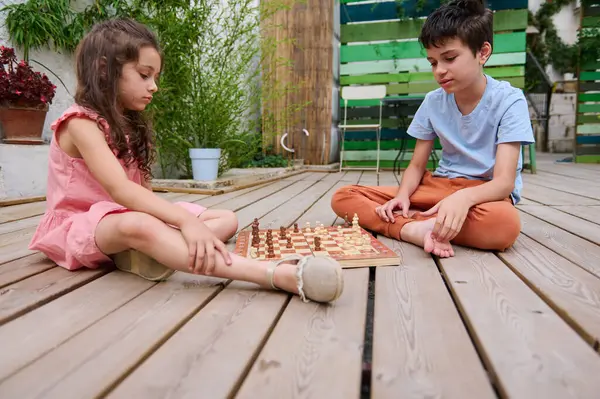 Stock image Two children engaged in a chess game outside on a wooden deck in a garden, fostering strategic thinking and concentration.