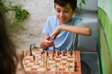 Neşeli bir çocuk bir arkadaşıyla açık havada satranç oynuyor, güneşli bir günün tadını çıkarıyor. Görüntü, satranç oyunundaki eğlence ve konsantrasyonu yakalar..