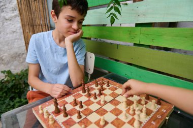 Açık havada satranç oynayan iki çocuk, strateji ve konsantrasyon gösteriyorlar. Bahçe ortamında yakalanmış, açık hava aktivitelerinin neşesini vurguluyor..