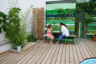 Anne ve kızı, canlı yeşillik ve renkli ahşap zemin ile sıcak bir verandada birlikte kaliteli zaman geçiriyorlar. Sahne sıcaklık, bağ ve açık hava rahatlığını temsil ediyor..