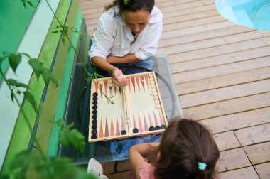 Anne ve kızı dışarıda ahşap bir güvertede tavla oynuyorlar. Canlı, doğal bir ortamda aile bağlarını ve boş zamanlarını ön plana çıkarıyorlar..