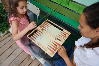 Bir anne ve kızı açık hava ortamında cam bir masada tavla oynuyorlar. Birlikte kaliteli bir aile zamanı geçiriyorlar..
