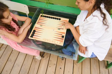 Bir anne ve kızı evde tavla oynamaktan zevk alır. Pembe elbiseli bir çocuk ve beyaz gömlekli bir kadın tahta bir masanın karşı tarafında oturuyorlar..