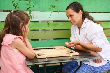 Anne ve kızı dışarıda masa oyunu oynayarak güzel vakit geçiriyorlar. Kadın onunla meşgul olurken çocuk düşünceli görünüyor..