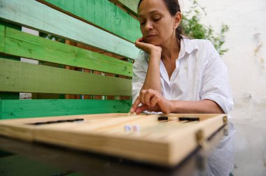 A woman sits outside in a garden playing backgammon. She is focused on the board game, enjoying a relaxing moment.