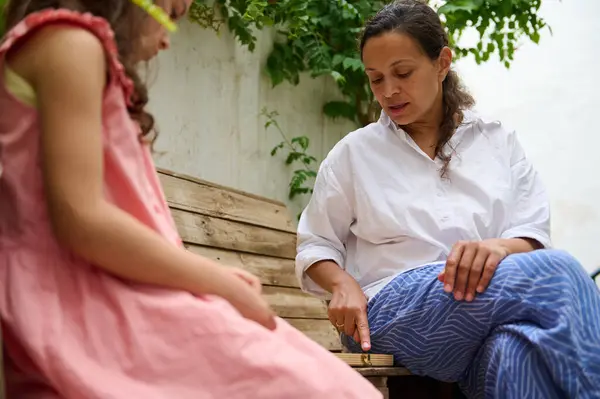 stock image A mother and daughter enjoy quality time together in their backyard, seated on a wooden bench. The mother engages the child, fostering a warm and nurturing environment.