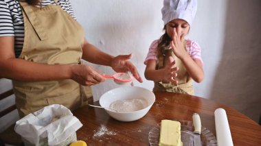 Önlüklü bir anne ve kızı sıcacık bir mutfakta birlikte yemek pişiriyorlar. Malzemeleri ölçüyorlar ve pişirme süreciyle eğleniyorlar, aile bağlarını ve aşçılık yaratıcılığını somutlaştırıyorlar..