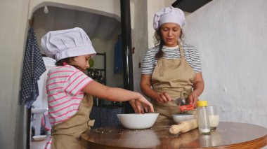 Anne ve kızı mutfakta birlikte yemek pişiriyor, önlük ve şef şapkası takıyor. Genç kız malzemeleri karıştırırken annesi de onları denetliyor..