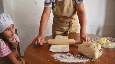 Bir anne ve kızı evde birlikte yemek pişirir. Annesi hamuru merdaneyle sararken kız da önlük ve aşçı şapkası takarak izliyor..