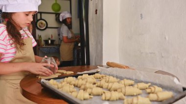 Genç bir kız ve annesi bir ev mutfağında birlikte hamur işi pişiriyorlar. Her ikisi de önlük ve şef şapkası takıyor, eğlenceli ve ilgi çekici bir yemek pişirme deneyimi yaratıyorlar..