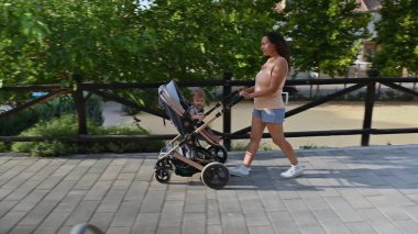 Genç anne çocuğuyla birlikte güneşli bir günde, yemyeşil ağaçlarla çevrili asfalt bir parkta yürüyor..