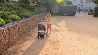 Genç bir anne, güneşli bir parkta bebek arabasıyla gezintiye çıkıyor. Arka planda yeşillik ve bir oyun alanı görülebilir.