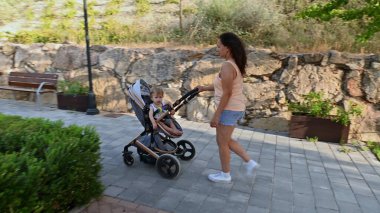 Çocuğuyla birlikte parkta bebek arabasını iten bir anne. Park, barışçıl ve aile odaklı bir gezinin altını çizen taş bir duvar ve yeşilliğe sahip..