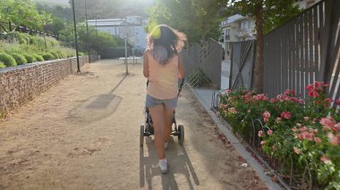 Parkta bebek arabasıyla yürüyen bir kadının arka görüntüsü. Doğal çevre ve çiçek açan güneşli bir gün. Huzurlu bir ortamda sakin bir şekilde dolaşmak..