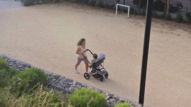 Bir anne parkta bebek arabasıyla koşarken, bir bebeği taşırken görülüyor. Gün açık ve açık hava aktiviteleri için mükemmel..