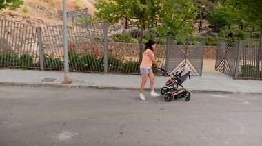 Gündelik kıyafetli genç bir kadın güneşli bir günde parkta bebek arabasını iterken koşuyor. Sahne fitness, annelik ve aktif bir yaşam tarzını betimliyor..