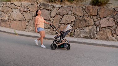Genç bir anne, arka planda taşralı bir duvar bulunan bir bebek arabasını şehir caddesinde gezdirirken gündelik bir yürüyüş yapmaktan zevk alır..