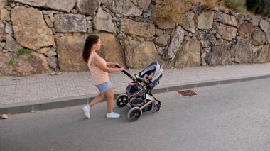 Bir annenin bebeğiyle birlikte arka planda taş bir duvarla sessiz bir cadde boyunca gezindiği görüntüsü..