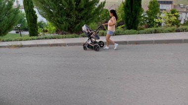 Güneşli bir günün tadını çıkaran genç bir anne huzurlu bir mahallede bebek arabasını itiyor. Sokaklar yemyeşil, huzurlu manzarayı güzelleştiriyor..