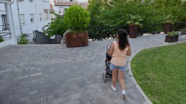 Güzelce döşenmiş bir şehir parkında bebek arabasını iten bir kadının arka görüntüsü. Arka planda yeşillik ve konut binaları.