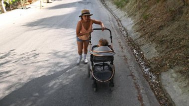 Bir anne bebeğini sessiz bir yolda yokuş yukarı itiyor. Sahne güneşli bir günde, sakin ve aktif bir açık hava atmosferi yaratıyor..