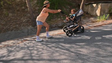 Enerjik anne, bebek arabasıyla ağaçlık bir yolda koşuyor. Sahne, aktif ebeveynlik ve açık hava sporunun özünü yakalıyor..