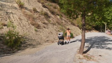 Doğayla çevrili dolambaçlı bir dağ yolunda bebek arabasını iten bir kadın. Güneşli bir gün ve açık hava bir rahatlama ve macera hissi veriyor..