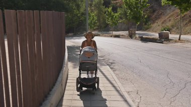 Bir kadın, sessiz bir banliyö caddesi boyunca bir bebek arabasıyla güneşli bir gündüz yürüyüşünün tadını çıkarıyor. Sahne, sükunet, rahatlama ve günlük rutin duyguları çağrıştırıyor..