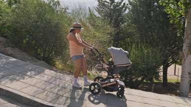 Anne dışarıda yürümekten zevk alıyor, güneşli bir günde yeşilliklerle çevrili bir parkta bebek arabasını yokuş yukarı itiyor..