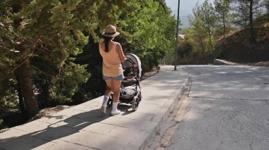 Bir anne bebek arabasını iterek manzaralı bir orman yolunda yürüyor. Aydınlık yaz günü, aile aktiviteleri için uygun huzurlu ve rahatlatıcı ortamı vurguluyor.