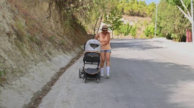 Güneşli bir günde huzurlu bir yürüyüş yapan bir kadın, doğayla çevrili sessiz bir yolda bebek arabasını itiyor. Sıradan giyinmiş ve şapka takıyor..