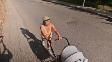 Yazın giyinmiş ve şapkalı bir kadın güneşli bir günde bebek arabasını tepeye iterken görülmüş. Banliyö ortamında sakin bir açık hava anını yakalamış..