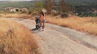 Şapkalı bir anne, güneşli bir günde, kuru otlar ve uzak evlerle çevrili, dolambaçlı kırsal bir yolda bebek arabasıyla yürüyor..