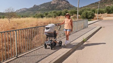 Yaz kıyafetleri içinde bir kadın manzaralı bir yolda bebek arabasıyla yürüyor, arka planda dağların ve yeşilliklerin olduğu kırsal bir bölgede güneşli bir günün tadını çıkarıyor..