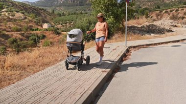 Açık, güneşli bir günde, kırsal kesimde, manzaralı tepeler ve yeşilliklerle bebe bebek arabasıyla yürüyen bir kadın. Annelik ve doğa yürüyüşü..