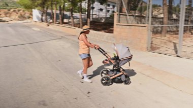 Bebek arabasıyla konut alanında koşan bir kadın. O günlük yaz kıyafetleri ve bir şapka giyiyor. Arka planda ağaçlar ve evler görülebilir..